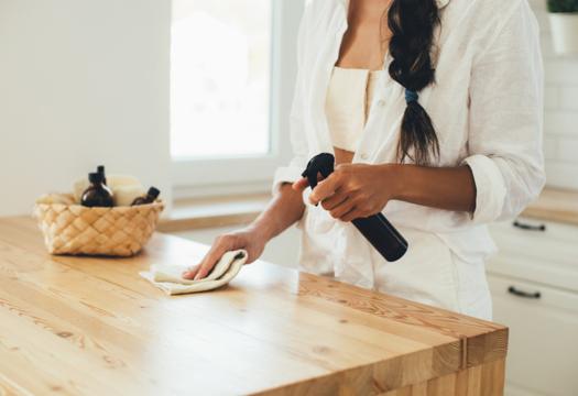 A young adult woman cleans her wooden kitchen island with a spray bottle and a rag. | Merry Maids